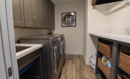 This full home renovation went from a complete gut to completely gorgeous. The laundry room features Mink Gray cabinetry from the Wellborn Premier series, under mount sink with quartz countertop, plenty of open shelving for baskets and storage, dedicated space for the homeowners pets and a cut out between rooms for laundry basket access. [TWD Project Location: Litchfield Park, AZ]