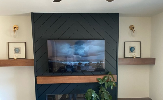 A custom media wall such as this is inclusive of the Herringbone shiplap wall painted in Black of Night, Caramel Maple cabinetry, mantle, floating shelves and added support for wall mounting the customers television and fireplace. [TWD Project Location: Phoenix, AZ]