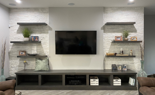 A custom media wall such as this inclusive of framing a pop-out, adding additional support for wall mounting the customers' television, electrical relocation, floating shelves and Wellborn cabinetry for additional storage and seating, as well as gorgeous decorative stone panels installed from floor to ceiling on each side of the television. [TWD Project Location: Phoenix, AZ]