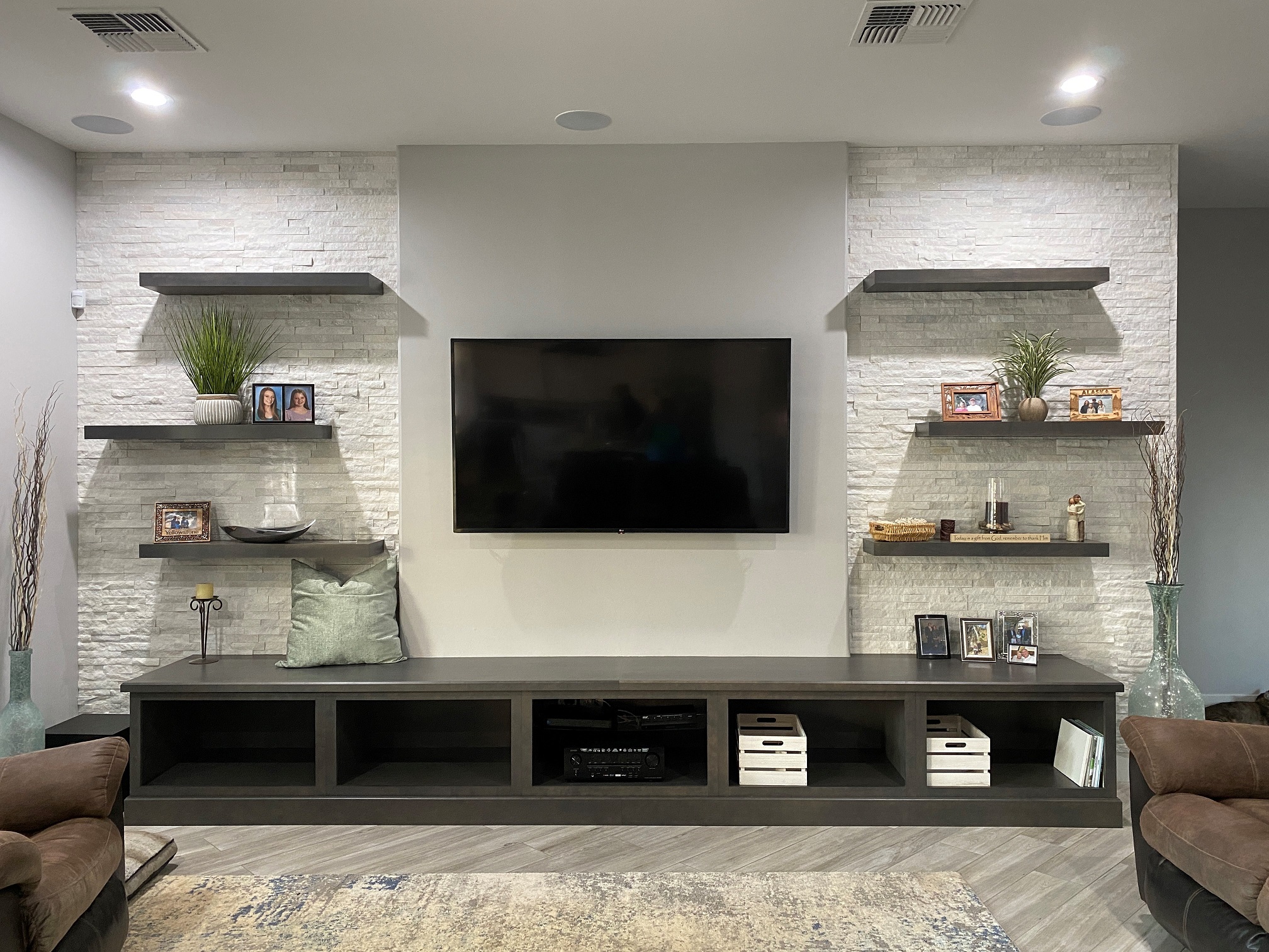 A custom media wall such as this inclusive of framing a pop-out, adding additional support for wall mounting the customers' television, electrical relocation, floating shelves and Wellborn cabinetry for additional storage and seating, as well as gorgeous decorative stone panels installed from floor to ceiling on each side of the television. [TWD Project Location: Phoenix, AZ]