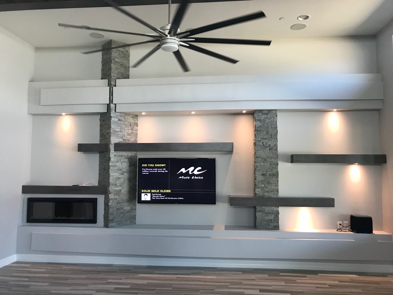 Custom media wall showcasing floating shelves in Maple Smoke finish, Sage Green ledger stone and plenty of dimmable lighting to set the mood of your room. [TWD Project Location: Peoria, AZ]