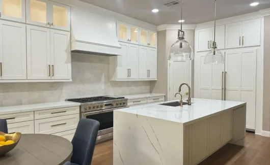 A modern kitchen featuring a spacious quartz countertop island with sleek white cabinetry, gold accents, and pendant lighting. The kitchen includes built-in appliances and a tiled backsplash, creating a clean and sophisticated aesthetic.