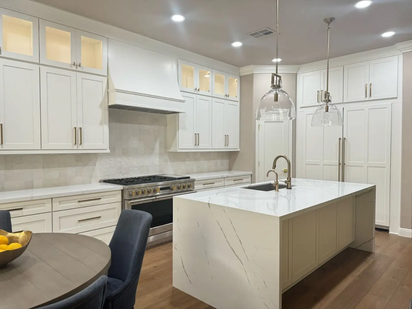 A modern kitchen featuring a spacious quartz countertop island with sleek white cabinetry, gold accents, and pendant lighting. The kitchen includes built-in appliances and a tiled backsplash, creating a clean and sophisticated aesthetic.