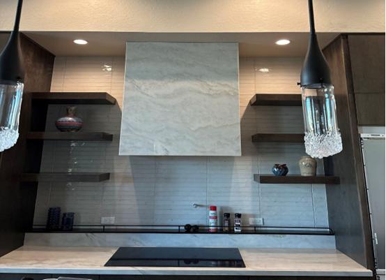 A modern kitchen featuring a sleek, custom marble range hood with clean lines. Floating dark wood shelves on either side display decorative pottery and functional kitchen items. The backsplash is tiled with subtle, glossy horizontal lines, and pendant lights with crystal accents hang above the space for an elegant touch.