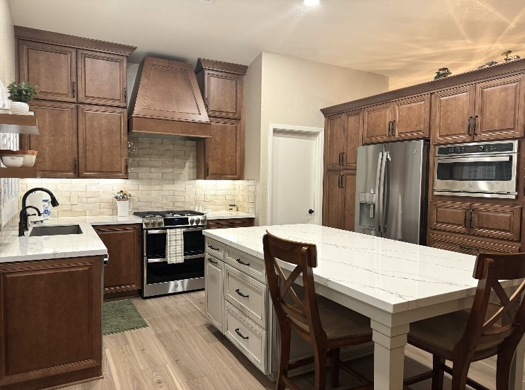 A beautifully remodeled kitchen featuring earthy and neutral tones. Warm wood cabinets complement the sleek white quartz island with veining, paired with stylish bar chairs. The kitchen boasts a statement wooden range hood, light beige brick backsplash, stainless steel appliances, and ample storage. Open shelving adds a modern touch, while the wood flooring ties the design together.