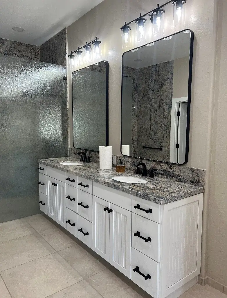 A luxurious bathroom with a double-sink vanity featuring granite countertops, white shaker cabinets, and framed mirrors under elegant light fixtures. The space is complemented by a sleek glass shower and soft neutral tones for a relaxing ambiance. Upgrade your bathroom for a touch of sophistication today.