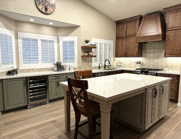 A remodeled kitchen featuring a spacious white quartz island with elegant veining and green cabinets with dark handles. The kitchen includes warm wood-toned cabinetry, a stylish range hood, and light brick-style backsplash. Large plantation shutters allow natural light to flood in, complementing the modern lighting fixtures. A wine cooler is built into the lower cabinetry, while wooden flooring ties the room together.