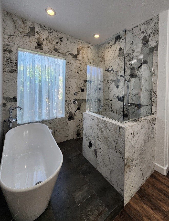 A luxurious bathroom featuring a freestanding white bathtub and a modern walk-in shower enclosed by glass. The walls are adorned with bold, dramatic marble, blending dark veining with light tones. A window with sheer curtains adds natural light, contrasting with dark floor tiles for a striking visual.