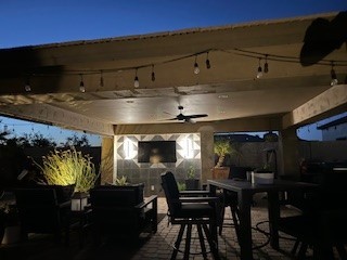 A cozy outdoor patio space illuminated at night. The covered area features string lights, a ceiling fan, and a mounted TV surrounded by sleek lighting fixtures. Comfortable seating and a dining table complete the inviting atmosphere, ideal for evening gatherings.