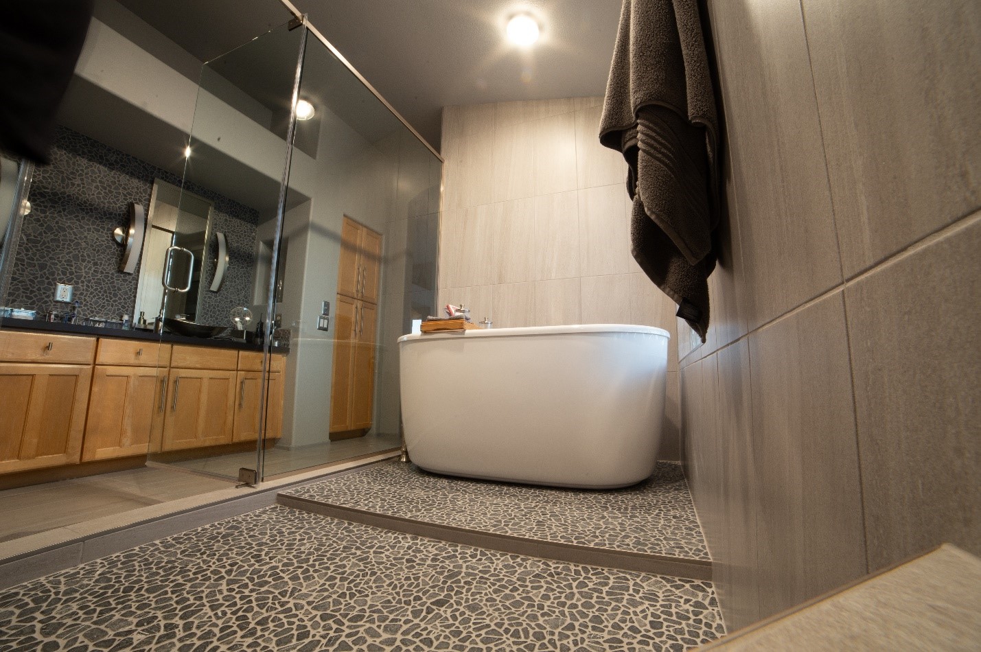 A luxurious bathroom featuring a freestanding white soaking tub and a glass-enclosed shower. The floor is covered in pebble-style tiles, adding texture and a spa-like feel. Light wood cabinetry complements the soft gray wall tiles, and modern lighting enhances the serene ambiance.