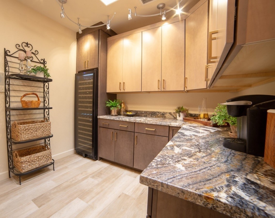 A beautifully designed walk-in pantry featuring elegant cabinets in neutral tones and a dramatic granite countertop with intricate veining. The room includes a built-in wine cooler, a stylish metal shelving unit with woven baskets, and warm track lighting that enhances the cozy yet modern ambiance. Light wood flooring completes the look.