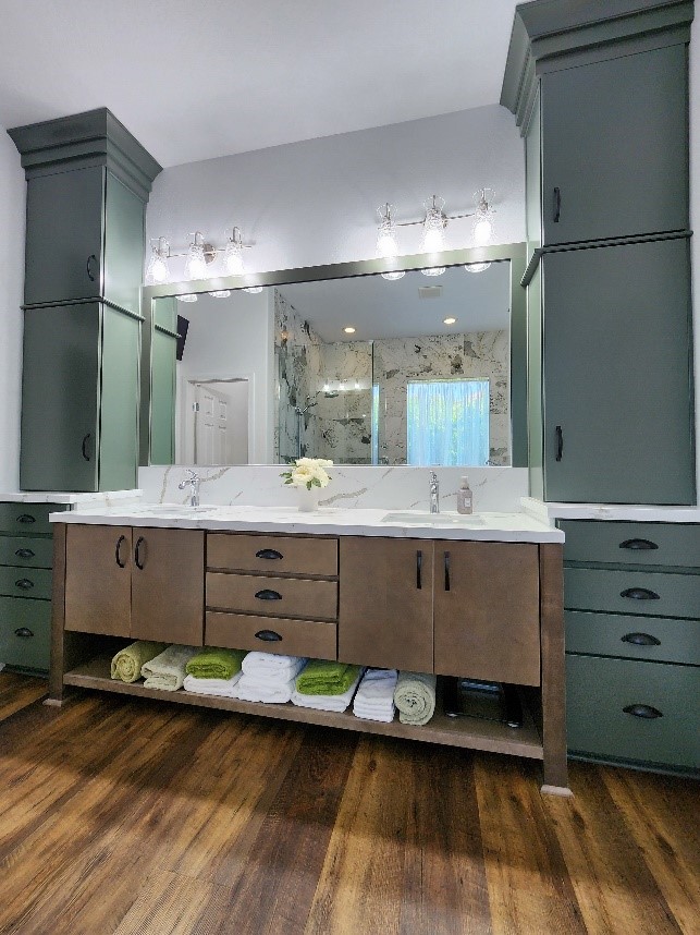 A contemporary bathroom with dark green cabinets, a white marble countertop, and a wood-toned vanity featuring open shelving for neatly folded towels. The space is complemented by modern light fixtures and a large mirror.