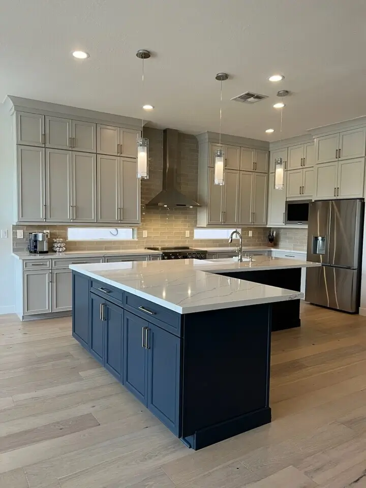 A modern kitchen featuring a large navy blue island with a white quartz countertop, light wood flooring, beige cabinetry, stainless steel appliances, and elegant pendant lighting.