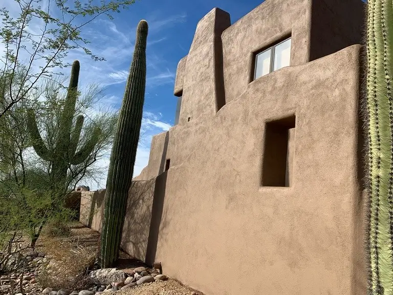 A stunning adobe-style home in the Arizona desert, surrounded by towering saguaro cacti and desert landscaping under a bright blue sky.