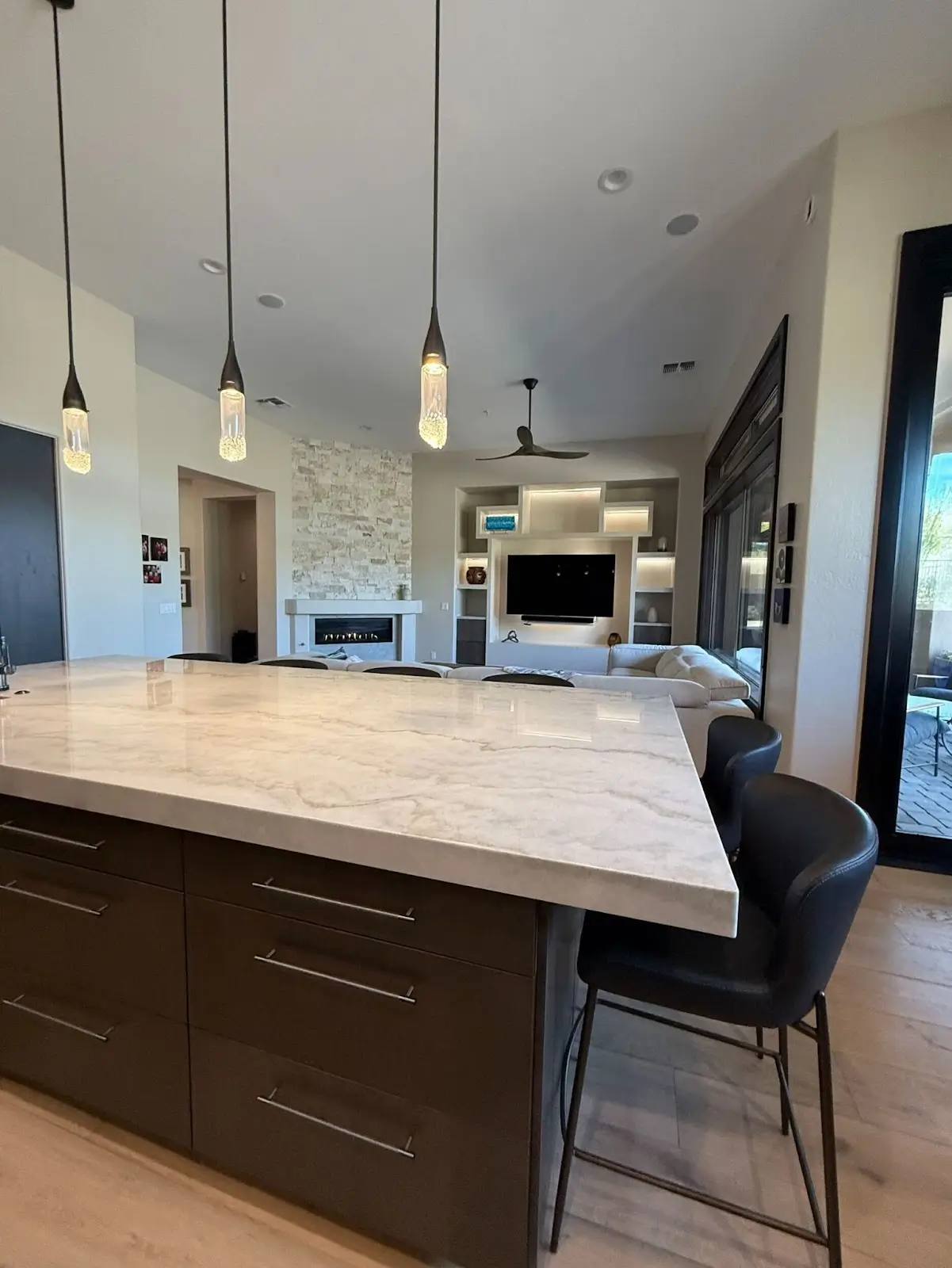 A modern kitchen featuring a large marble island with dark cabinetry, sleek pendant lighting, and an open-concept layout leading into a cozy living room with a built-in media wall and fireplace.