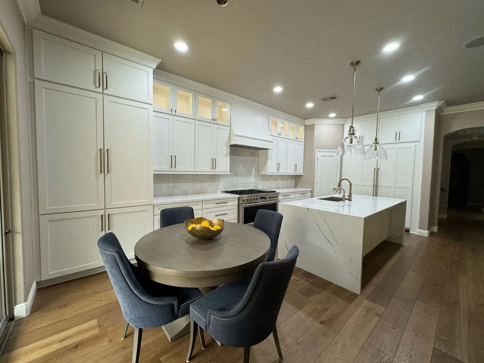 Modern kitchen interior featuring slip-resistant wood-look flooring, white cabinetry, a marble island, and a round dining table with navy upholstered chairs.